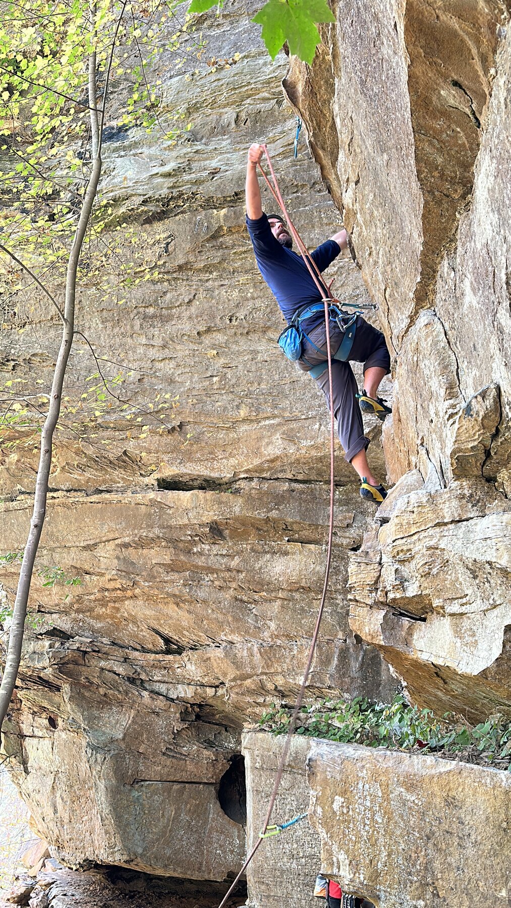 Climber pulling rope to clip a quickdraw on an overhanging Red River Gorge sport route, gear sling visible on harness